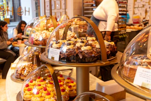 A bakery display counter showing a variety of colorful and elaborate pastries under glass domes. Different types of cakes, cupcakes, and tarts are neatly arranged. Customers are seen in the background, with one individual standing behind the counter and another seated, probably enjoying some pastries. The setting appears to be warm and inviting with a cozy atmosphere.