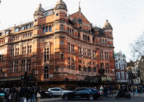 A grand, historic red-brick building with intricate architectural details and multiple arched windows stands prominently at a busy street corner. People are seen walking on the sidewalk, and several cars and motorbikes are passing by. The facade displays a sign for a Harry Potter play.