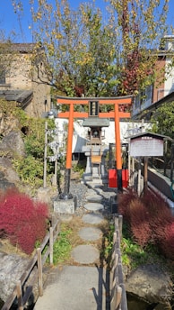 A picturesque scene features a traditional Japanese torii gate painted in vibrant orange and black, leading to a small shrine. The stone pathway is flanked by neatly trimmed bushes and trees with autumn foliage, adding bursts of red and green. This idyllic setting is surrounded by rustic buildings, enhancing its serene and cultural ambiance.