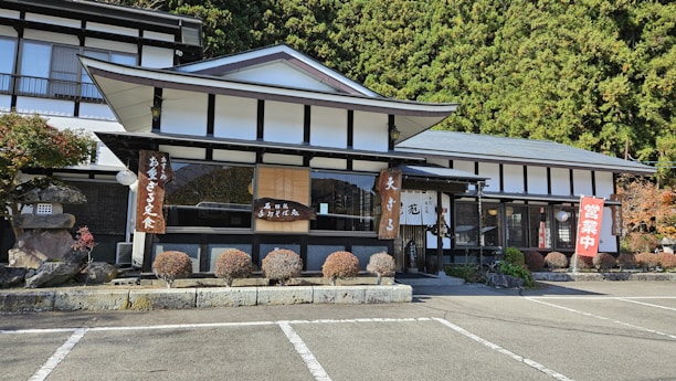 A traditional Japanese building with a black and white exterior is nestled among dense greenery. The structure features a sloping roof and several signs with Japanese characters hang from the eaves. Well-maintained shrubs line the front of the building, and there is a parking area in the foreground.