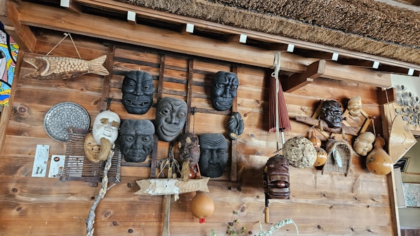 A wooden wall displays an assortment of traditional masks and handcrafted items. Several masks with expressive faces are arranged alongside woven gourds and decorative objects, including a wooden fish sculpture and an ornamental fan. The design reflects a rustic, cultural collection.