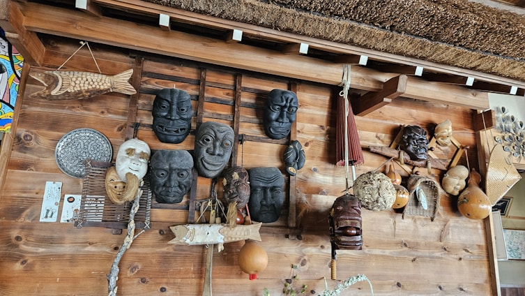 A wooden wall displays an assortment of traditional masks and handcrafted items. Several masks with expressive faces are arranged alongside woven gourds and decorative objects, including a wooden fish sculpture and an ornamental fan. The design reflects a rustic, cultural collection.