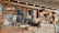 A wooden wall displays an assortment of traditional masks and handcrafted items. Several masks with expressive faces are arranged alongside woven gourds and decorative objects, including a wooden fish sculpture and an ornamental fan. The design reflects a rustic, cultural collection.