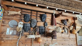 A wooden wall displays an assortment of traditional masks and handcrafted items. Several masks with expressive faces are arranged alongside woven gourds and decorative objects, including a wooden fish sculpture and an ornamental fan. The design reflects a rustic, cultural collection.