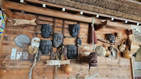 A wooden wall displays an assortment of traditional masks and handcrafted items. Several masks with expressive faces are arranged alongside woven gourds and decorative objects, including a wooden fish sculpture and an ornamental fan. The design reflects a rustic, cultural collection.