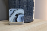 A small, square electronic device with a swirling, marble-like pattern in blue, gray, and black rests on a light wooden surface. It is placed next to a tall, textured ceramic vase with a geometric pattern, colored in a deep blue. The setup appears minimalistic and aesthetically coordinated, suggesting a modern and stylish interior arrangement.