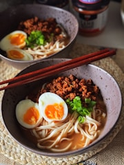 A bowl of ramen with noodles, two halves of a soft-boiled egg with orange yolks, minced meat, and chopped green onions. Wooden chopsticks rest on the edge of the bowl. A second bowl is partially visible in the background, alongside some condiment jars.