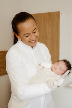 A person is joyfully holding a sleeping newborn dressed in warm, light-colored clothing. The person is dressed in a white outfit and is gazing lovingly at the baby. The background is neutral, featuring a hint of wooden texture from a piece of furniture.