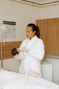 A woman dressed in a white lab coat is holding a newborn baby in a hospital room. The baby is wrapped in a light-colored blanket and has dark hair. In the background, there is medical equipment, including an IV stand and a hospital bed.