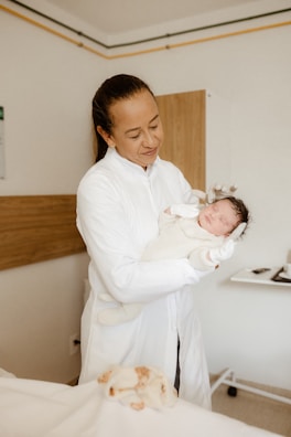 A person wearing a white medical coat gently holding a newborn baby, dressed in a warm, light-colored outfit. The setting appears to be a hospital room with beige and white tones, featuring a wall with a wooden panel and a small table in the background.