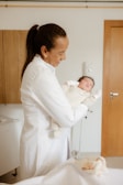 A person wearing a white coat holds a newborn baby wrapped in a cream-colored outfit in a warmly lit room with wooden elements. The individual appears to be smiling gently at the baby.