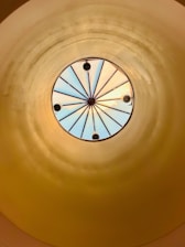 A circular skylight with a radial design featuring multiple glass sections separated by metal beams. The light entering through the skylight creates a play of colors and reflections on the surrounding dome-like structure, which is painted in a warm, golden hue.