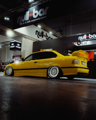 A bright yellow car with sleek, sporty features is prominently displayed against the backdrop of an exhibition booth. The car has custom rims and a prominent spoiler, surrounded by branding for 'null-bar' which specializes in air and static suspension parts. The setting is a well-lit indoor showroom or auto exhibition.