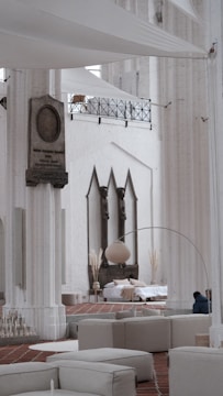 A spacious interior with tall white columns and arches, featuring contemporary minimalist decor. There is a seating area with light-colored couches and a large floor lamp. In the background, a bed is set up with cushions and decorative plants, situated against a wall with three pointed arches. The architectural details suggest a blend of modern and historical elements.