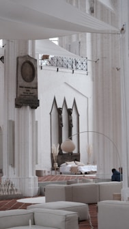 A spacious interior with tall white columns and arches, featuring contemporary minimalist decor. There is a seating area with light-colored couches and a large floor lamp. In the background, a bed is set up with cushions and decorative plants, situated against a wall with three pointed arches. The architectural details suggest a blend of modern and historical elements.