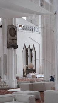 A spacious interior with tall white columns and arches, featuring contemporary minimalist decor. There is a seating area with light-colored couches and a large floor lamp. In the background, a bed is set up with cushions and decorative plants, situated against a wall with three pointed arches. The architectural details suggest a blend of modern and historical elements.