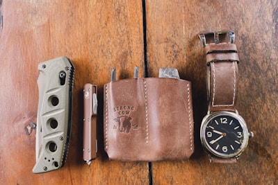 A rustic wooden surface features a collection of everyday carry items. A folding knife with a textured handle, a compact brown pocket tool, a leather pouch embossed with 'Strong Cow,' and a stylish wristwatch with a brown leather strap are neatly arranged in a row.