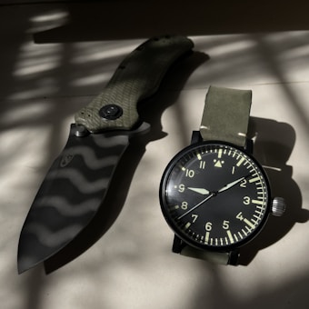 A folding knife with a textured handle and a camouflage-patterned blade is placed next to a wristwatch with a green strap and black face. Light and shadow patterns fall across both objects, creating an industrial and rugged feel.
