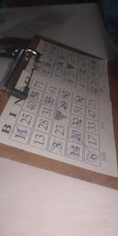 A bingo card is filled with numbers in a five-by-five grid, with several numbers crossed out using a blue pen. The grid is labeled with the letters B, I, N, G, O at the top. The card is clipped to a wooden clipboard, resting on a white, textured surface in a dimly lit area.