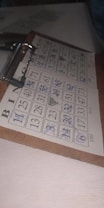 A bingo card is filled with numbers in a five-by-five grid, with several numbers crossed out using a blue pen. The grid is labeled with the letters B, I, N, G, O at the top. The card is clipped to a wooden clipboard, resting on a white, textured surface in a dimly lit area.