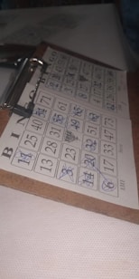 A bingo card is filled with numbers in a five-by-five grid, with several numbers crossed out using a blue pen. The grid is labeled with the letters B, I, N, G, O at the top. The card is clipped to a wooden clipboard, resting on a white, textured surface in a dimly lit area.