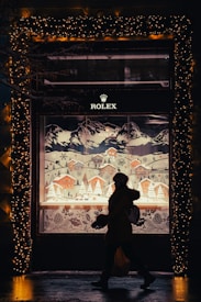 A person walking in front of a storefront display featuring a winter village scene with snow-covered houses and trees. The display is framed with sparkling holiday lights, creating a festive ambiance. A prominent brand logo is situated above the display.