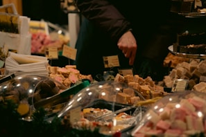 A variety of fudge and sweets are displayed at a market stall. The hand of a person is seen selecting or pointing at a piece. Labels are visible among the confections, offering a wide selection of flavors. The setup is cozy and warm, perfect for a festive or holiday market atmosphere.