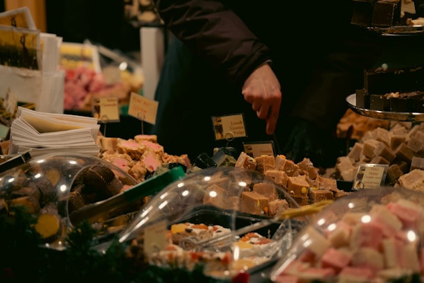 A variety of fudge and sweets are displayed at a market stall. The hand of a person is seen selecting or pointing at a piece. Labels are visible among the confections, offering a wide selection of flavors. The setup is cozy and warm, perfect for a festive or holiday market atmosphere.