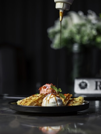 A plate of waffles is served with two scoops of ice cream, one strawberry and one vanilla, drizzled with a caramel sauce. Fresh mint leaves garnish the dish. The background is out of focus, highlighting the dessert as the main subject.