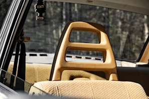 A vintage car interior with tan upholstery and a unique headrest design featuring cutouts. The rear window provides a view of a blurred, wooded background. A black air freshener in the shape of a tree hangs from the rearview mirror.