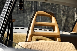 A vintage car interior with tan upholstery and a unique headrest design featuring cutouts. The rear window provides a view of a blurred, wooded background. A black air freshener in the shape of a tree hangs from the rearview mirror.