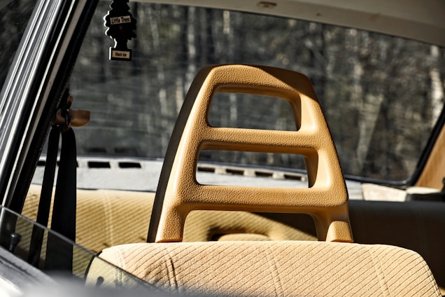 A vintage car interior with tan upholstery and a unique headrest design featuring cutouts. The rear window provides a view of a blurred, wooded background. A black air freshener in the shape of a tree hangs from the rearview mirror.