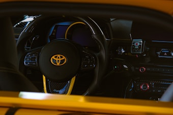 A close-up view of a car's interior featuring a steering wheel with the Toyota logo prominently displayed. The dashboard includes various controls and buttons, and there is a digital display just behind the steering wheel. The materials have a carbon fiber finish, highlighted by yellow accents around the steering wheel and dashboard.