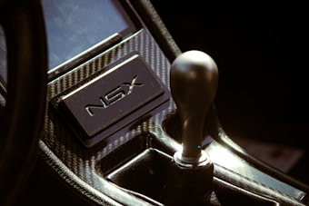 A close-up view of a car's interior featuring a gear shift knob and a plaque with the letters NSX, set in a carbon fiber console.