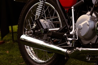 A close-up view of a motorcycle's mechanical components, including the exhaust pipe, wheel, tire, shock absorber, and engine parts. The image showcases metallic and chrome surfaces with a shiny appearance, highlighting the intricate design and engineering of the motorcycle.