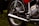 A close-up view of a motorcycle's mechanical components, including the exhaust pipe, wheel, tire, shock absorber, and engine parts. The image showcases metallic and chrome surfaces with a shiny appearance, highlighting the intricate design and engineering of the motorcycle.
