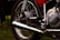 A close-up view of a motorcycle's mechanical components, including the exhaust pipe, wheel, tire, shock absorber, and engine parts. The image showcases metallic and chrome surfaces with a shiny appearance, highlighting the intricate design and engineering of the motorcycle.