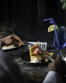 A person is using a fork and knife to cut into a dessert consisting of waffles with scoops of ice cream, garnished with mint and drizzled with syrup. A glass of blue drink with a straw and lemon slice is placed on the table next to a table number sign showing the number 9.