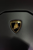 A close-up view of a Lamborghini emblem featuring a golden bull on a black shield, set against a matte dark surface.