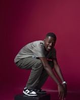 A person squats on a platform against a solid maroon background. They are wearing a gray shirt, olive green pants, and black and white sneakers. A shiny watch is visible on their left wrist.