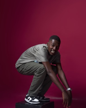 A person squats on a platform against a solid maroon background. They are wearing a gray shirt, olive green pants, and black and white sneakers. A shiny watch is visible on their left wrist.