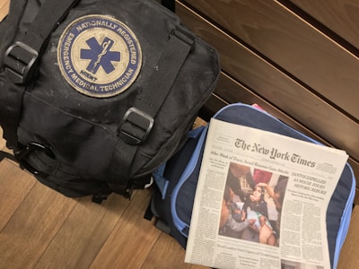 A black bag with a patch reading 'Nationally Registered Emergency Medical Technician' is placed next to a wooden surface. Beside it, a copy of The New York Times newspaper is laid on top of another bag, possibly blue. The newspaper article headline addresses the resumption of attacks in Gaza after a truce, and the image on the front shows a group of people attending to an injured individual.