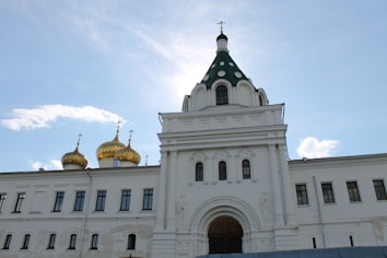 A large white Orthodox church with ornate architecture is highlighted against a clear blue sky. The building features distinct golden domes topped with crosses and a central tower with a green, patterned roof.