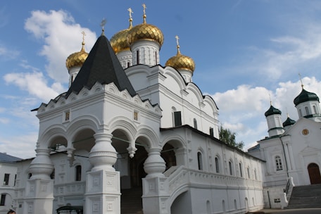 A white Orthodox church features multiple golden domes with crosses and elaborately detailed arches at the entrance. The building's architecture includes ornate carvings and pilasters, set against a bright blue sky with a few scattered clouds.