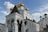 A white Orthodox church features multiple golden domes with crosses and elaborately detailed arches at the entrance. The building's architecture includes ornate carvings and pilasters, set against a bright blue sky with a few scattered clouds.