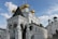 A white Orthodox church features multiple golden domes with crosses and elaborately detailed arches at the entrance. The building's architecture includes ornate carvings and pilasters, set against a bright blue sky with a few scattered clouds.