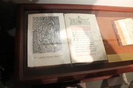 A display case containing an open, ornate old book with intricate illustrations and faded text. The pages feature elaborate designs and decorative elements typical of historical manuscripts. The item appears to be an ancient manuscript under protective glass, suggesting a museum or library setting.