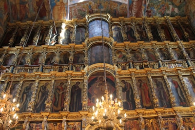 A richly decorated, multi-tiered iconostasis adorned with numerous religious paintings and ornate gold detailing. Brightly lit chandeliers cast a warm glow across the opulent structure, which features a combination of biblical figures and intricate carvings.