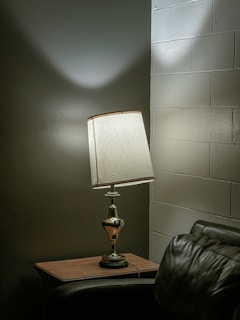 A dimly lit corner of a room featuring a vintage table lamp with a white lampshade placed on a wooden side table. The soft light casts distinct shadows on the dark textured wall and the adjacent white brick wall. Part of a dark leather sofa is visible in the foreground, adding to the cozy setting.