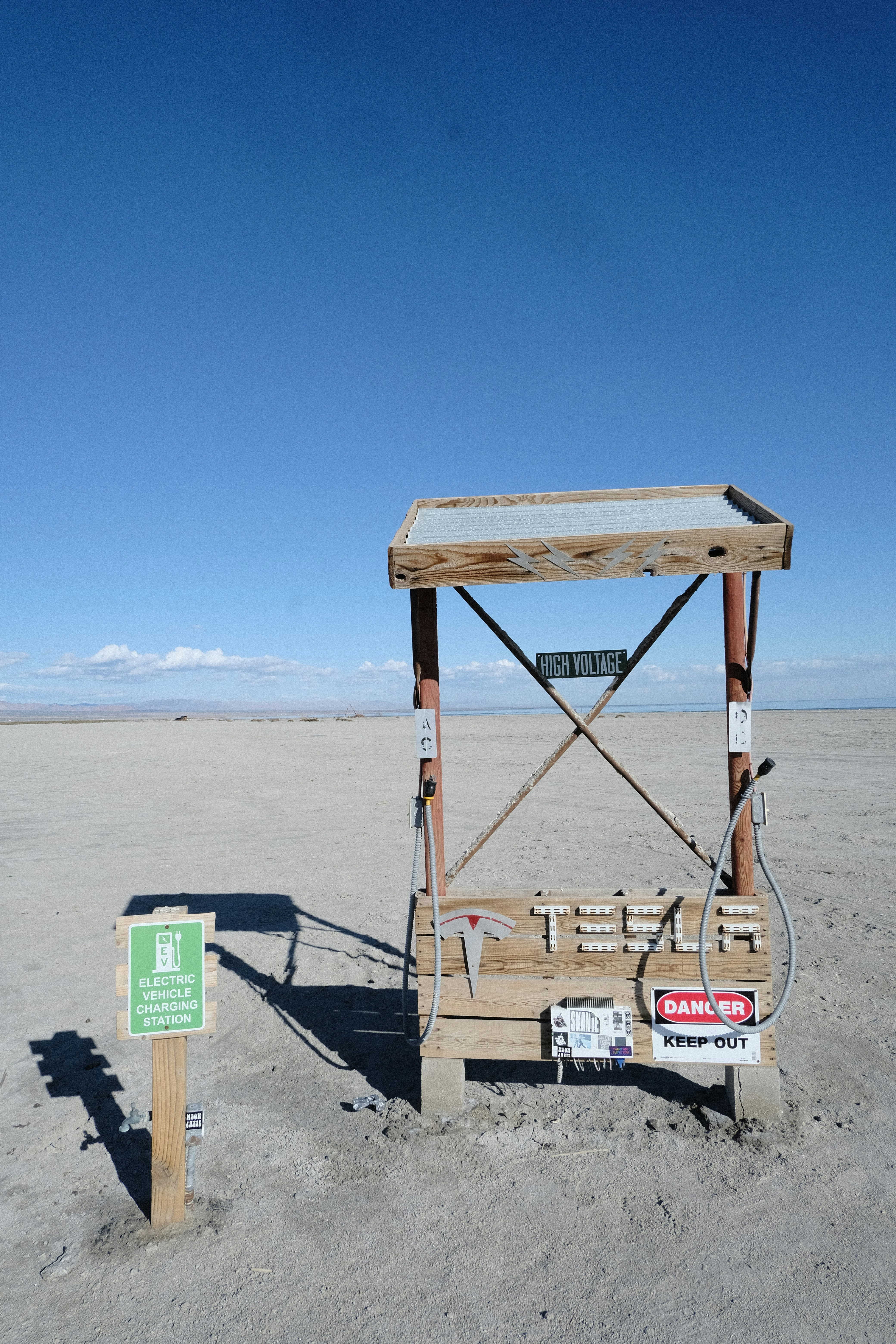 A minimalist electric vehicle charging station is situated in a barren, sandy landscape with a bright blue sky overhead. The charging station is marked by various warning signs, including 'High Voltage' and 'Danger Keep Out.' Nearby, a small sign indicates it's an electric vehicle charging point.